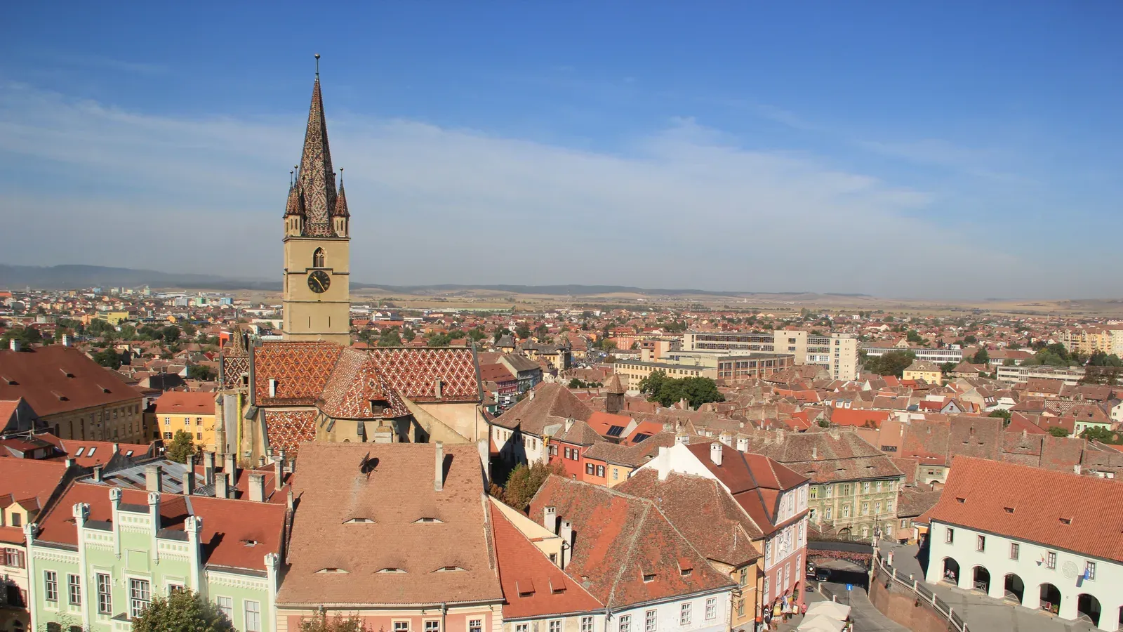 Red-tiled roofs of Sibiu's old town from above, with the spire of the Lutheran Cathedral and the Transylvanian plain beyond, Romania